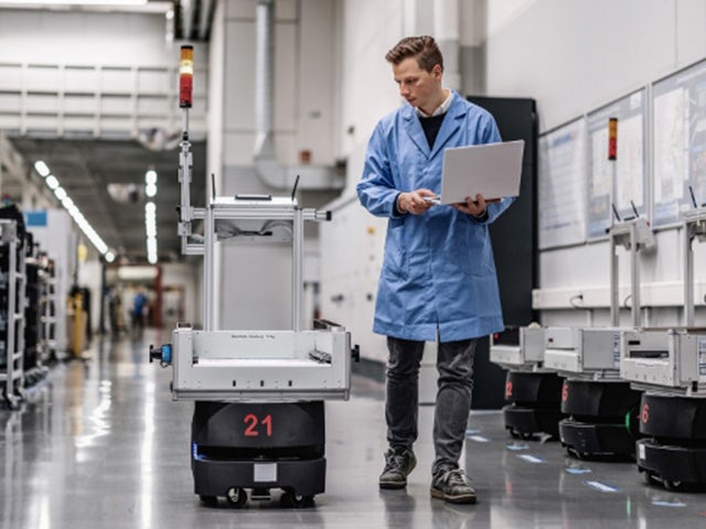 A man holding a laptop walks next to a mobile robot on a factory floor