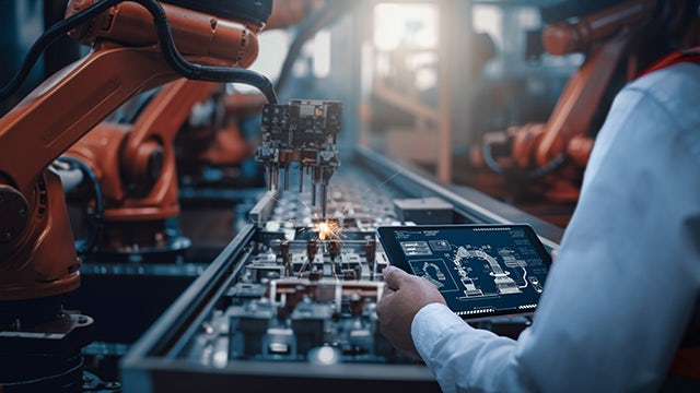 A person looks at a computer tablet while a robotic arm assembles parts on an assembly line