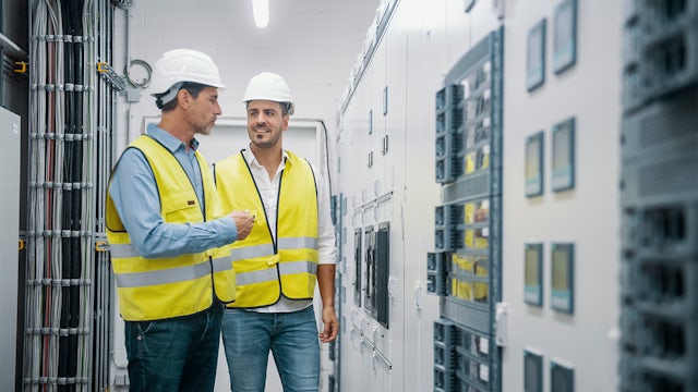 Two men in hi-vis jackets and white hard hats converse in an industrial setting.