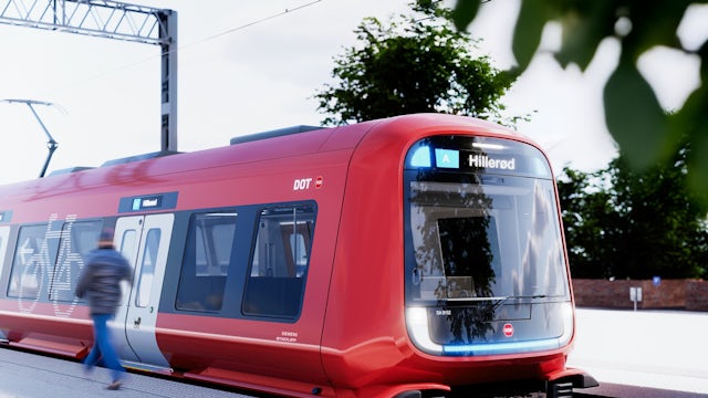 Red modern commuter train stopped at a station platform, with large front windows displaying the destination “Hillerød”; overhead power lines run above the track, and a blurred pedestrian walks past while trees and station structures appear in the background.