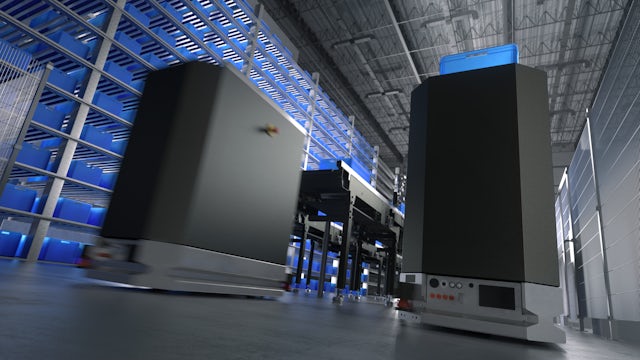 Two automated guided vehicles (AGVs) with tall black bodies and a blue bin on top of one, move across a warehouse floor with racks of blue storage bins in the background, illustrating automated logistics.