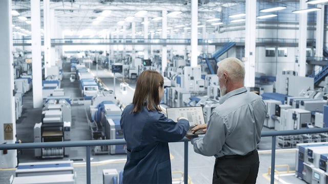 A male engineer and a female scientist using a laptop and talking in a factory facility.
