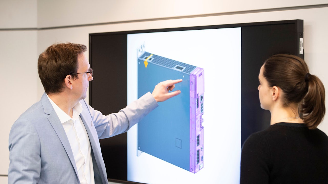 A Siemens expert points out a Digital Twin of a product on a screen to a colleague.</br>