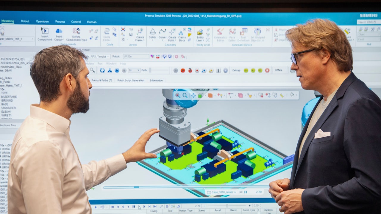 A Siemens expert points out a digital twin of a PCB production process on a screen to a colleague.</br>