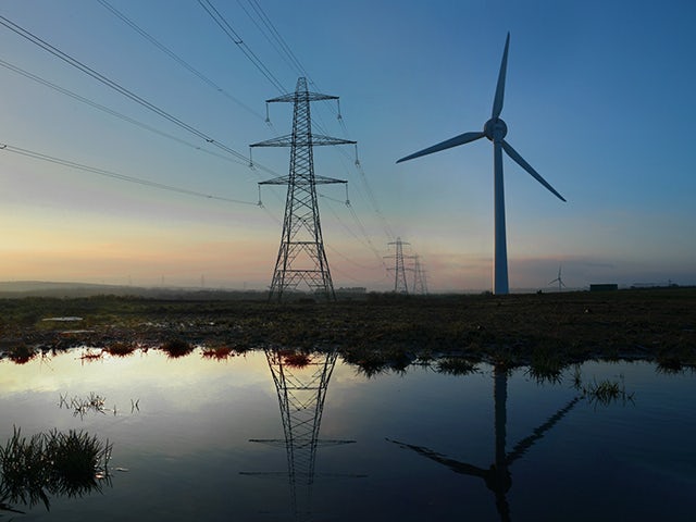 A large wind turbine stands tall against a twilight sky, with its reflection visible in a puddle of water in the foreground.