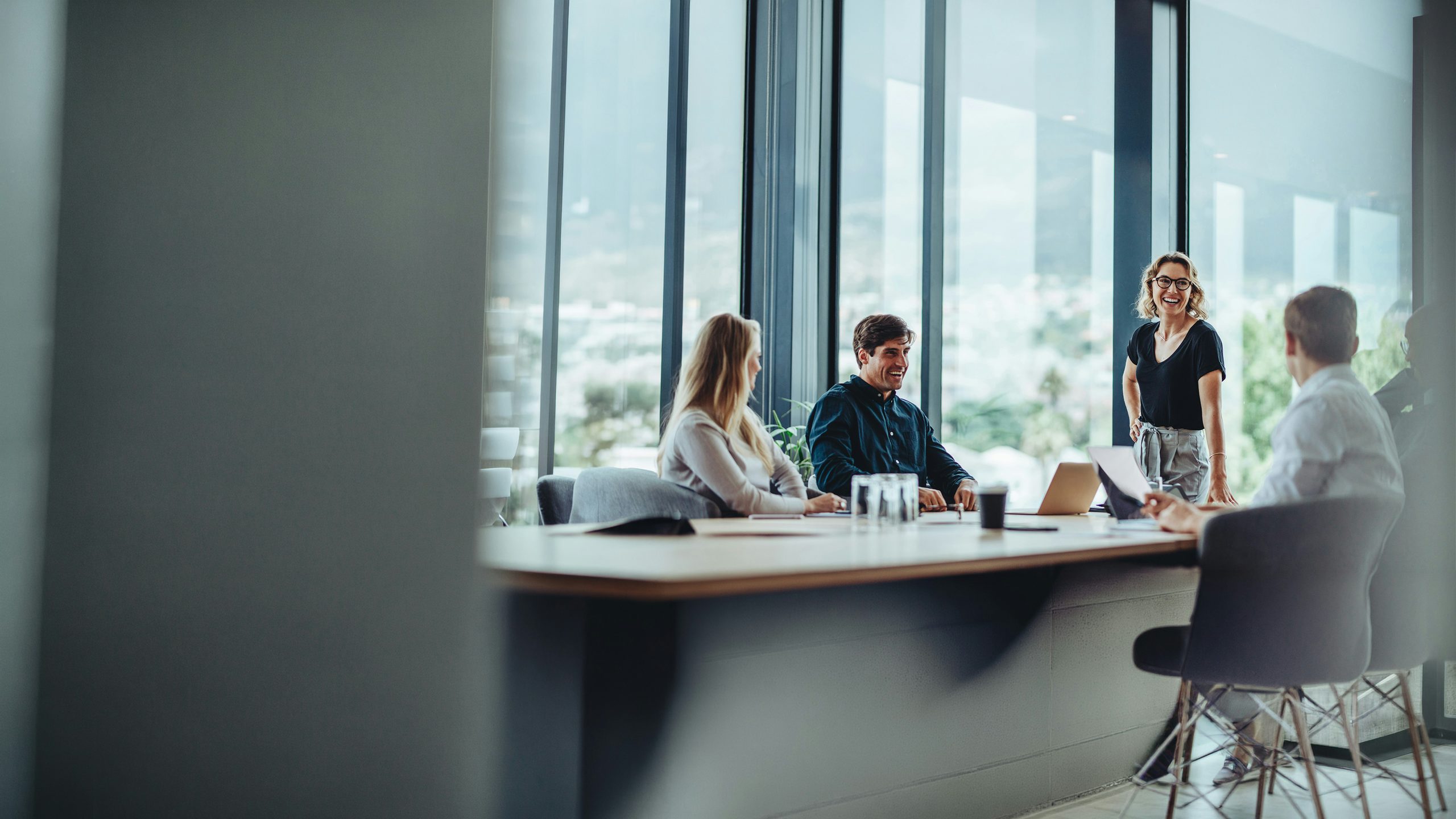 Business team meeting in a modern office, with a woman standing and speaking while colleagues sit around a conference table with laptops and documents.