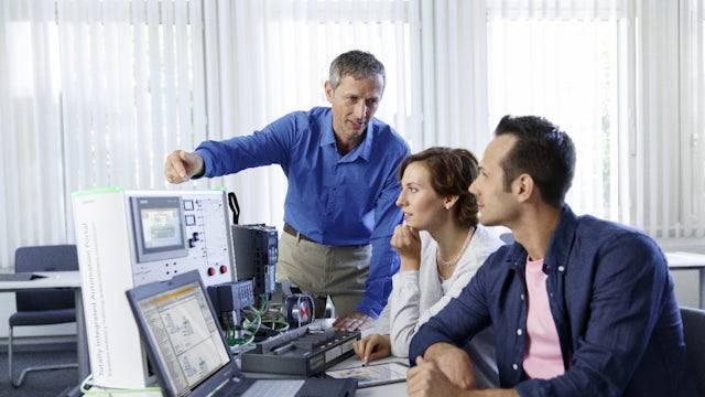 Three adults, two seated and one standing, are gathered around a table with a laptop and industrial control equipment, with the standing man pointing at a screen as the others observe.