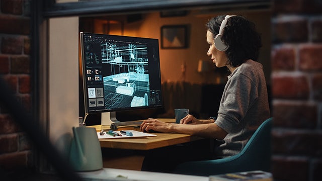 A woman wearing headphones sits in front of a computer running a design program