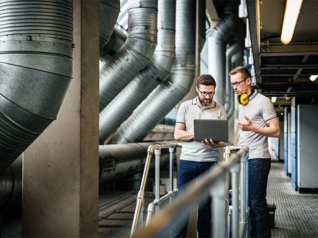 Two engineers with a laptop standing in a large printery and discussing latest production data.