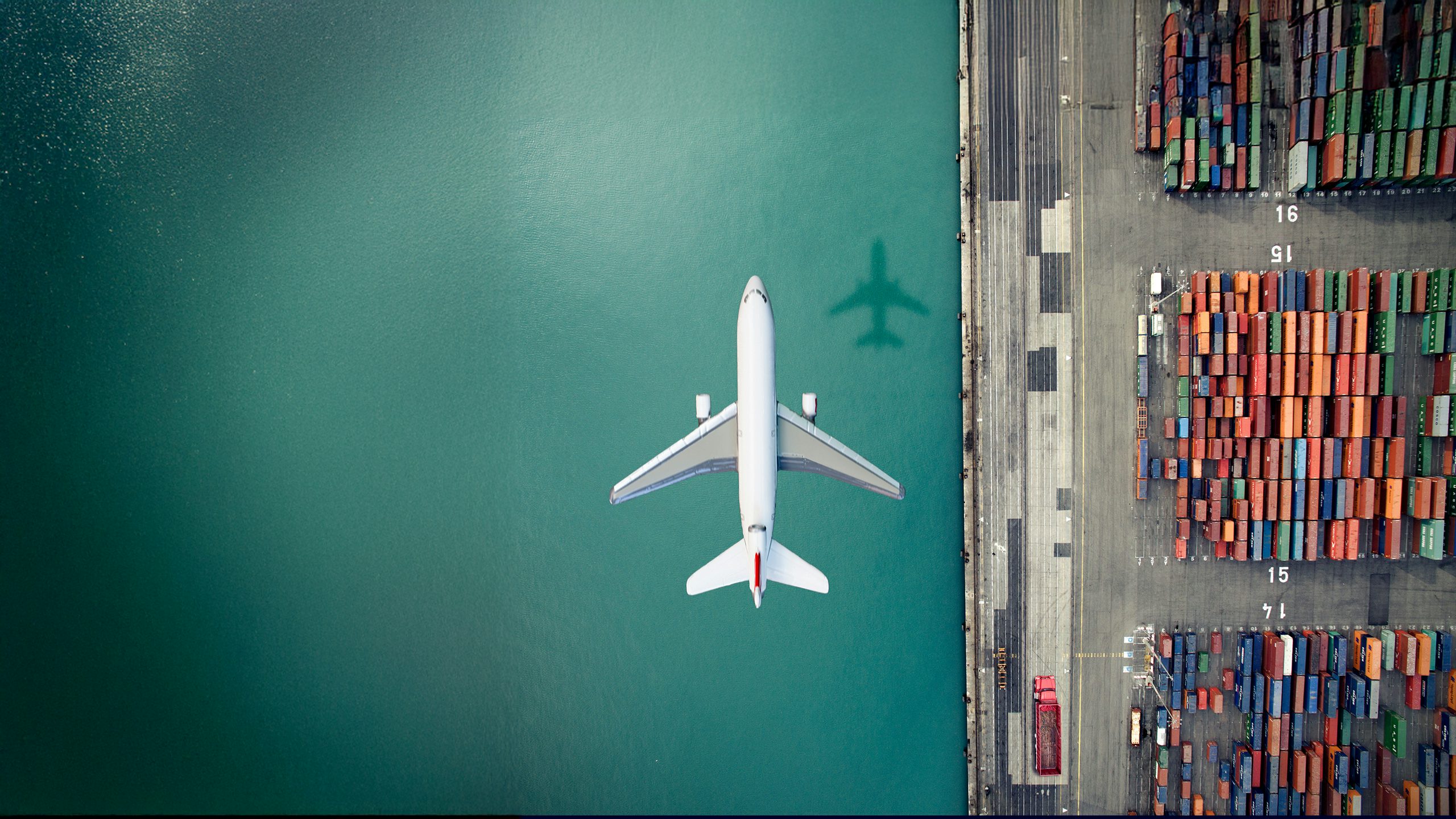 A plane flying over cargo containers on a body of water with a blue sky in the background.