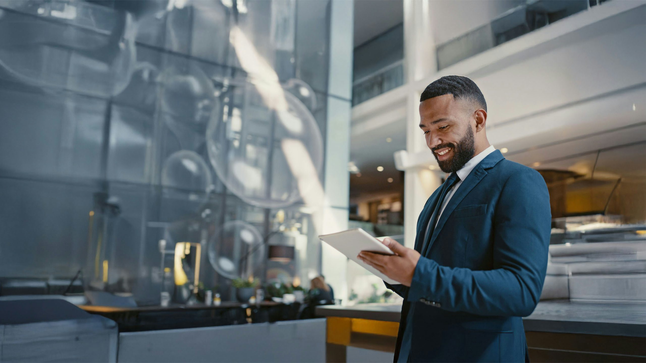 A smiling businessman in a dark blue suit uses a digital tablet while standing in a modern, well-lit corporate office lobby.