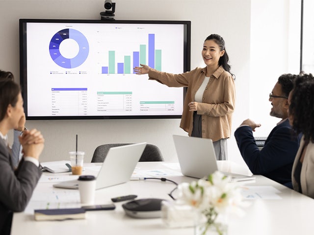 Woman speaking to a room pointing at a white board