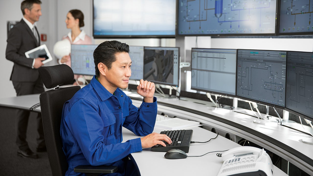 A control room with multiple monitors and a person standing in front of a desk.