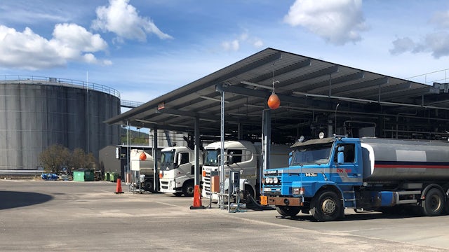 A truck is parked at a terminal with a large tank farm in the background.